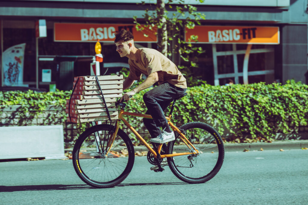 A man is riding a bicycle fast with ten pizza boxes packed on a front rack.