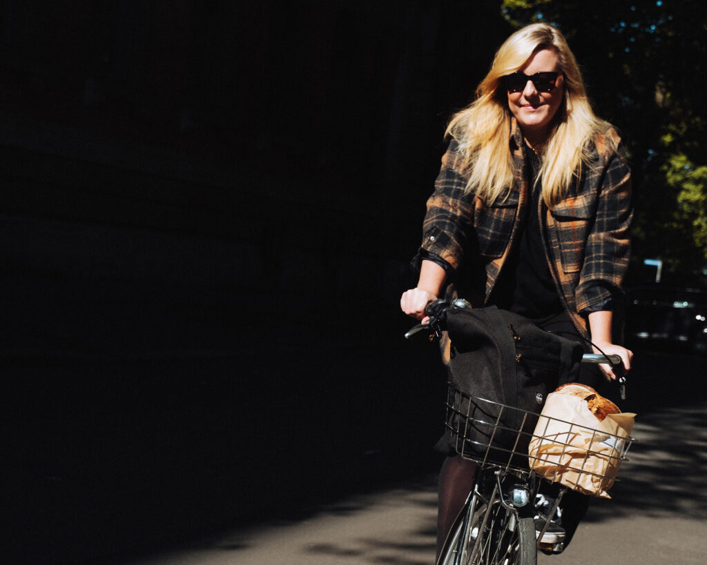 A women riding a bicycle with a backpack and bread packed in a bicycle basket.